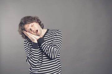 Young man in striped black and white sweater wants to sleep. Put his head in his hands and closed eyes while yawning. Standing on the grey background. Hands under his cheek. Insomnia concept