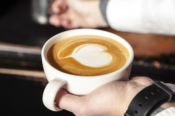 A professional Barista holds a Cup of coffee, making a beautiful heart shape latte art. Hot art latte coffee with heart shape in white Cup