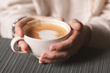 Close-up of a white Cup of hot latte art coffee with a heart shape in the hands of a young girl. Woman wearing warm winter knitted white sweater. Toned. Coffee Girl Concept Relaxation Coffee Mug.