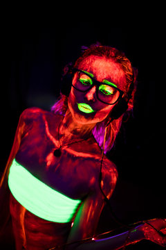 Portrait of a beautiful young sexy girl in headphones with ultraviolet paint on her body. Pretty woman with glowing bodyart in black lamp light