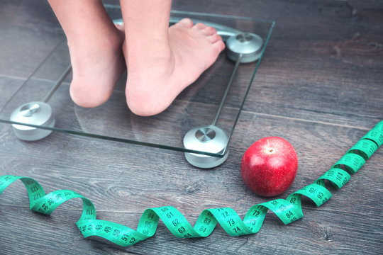 Green Measure Tape, Glass Weighing Scale, Red Apple And Feet On A Scale On A Rustic Surface. Weight Management And Healthy Wellbeing Concept