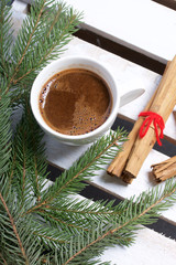 A cup of coffee and Cinnamon sticks next to the fir branches. On a wooden box.