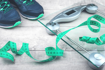 Fototapeta na wymiar Green measure tape, glass weighing scale, running shoes on a rustic surface. Weight management and healthy wellbeing concept