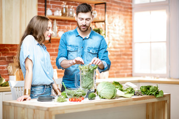 Young couple of vegetarians in blue shirts making smoothie with fresh green products on the kitchen at home