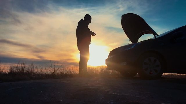 Silhouette man is checking engine bay of the broken down car while sunset.