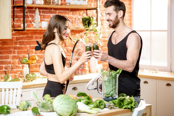 Young sports couple having a snack with vegetarian food drinking smoothie standing with rackets on the kitchen at home