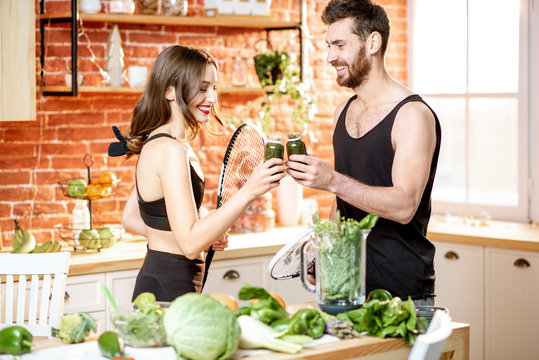 Young sports couple having a snack with vegetarian food drinking smoothie standing with rackets on the kitchen at home
