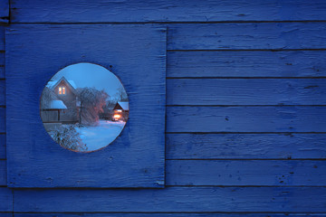 Fragment of a wooden fence with a round window. In the window is a beautiful winter night view of the village house. Toning