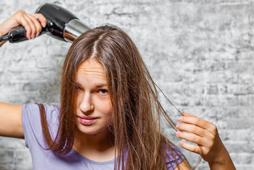 Fototapeta premium portrait of young teenager brunette girl with long hair dries hair with electrical hair dryer on gray wall background