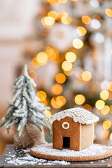 Gingerbread house on wooden table. Defocused garland lights on background. Christmas tree and Holiday mood. Morning in the bright living room.