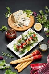 Pear with jerky meat, blue cheese and a mix of salad on a dark board. Next to olive oil, bread sticks with ham, lemon. Blurred background. Mediterranean cuisine
