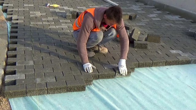 Construction worker laying interlocking paving concrete onto sheet nonwoven bedding sand and fitting them into place.