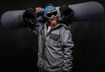 Brutal man with a red beard wearing a full equipment holding a snowboard on his shoulder, isolated on a dark textured background.