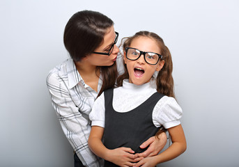 Happy mother whispering the secret to her excite kid in fashion glasses with opened mouth on empty copy space