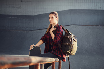 A young student girl with a backpack wearing a checkered shirt holding a skateboard while sitting on a grind rail in skatepark indoors. © Fxquadro