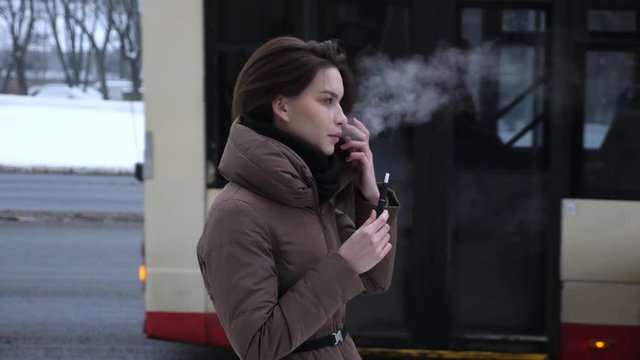 young beautiful woman smoking iqos outside cold winter, buss, city background 