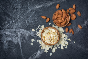 Top view of almond slices and whole nut in wooden bowl as ingredient for confectionery on black marble background with copy space
