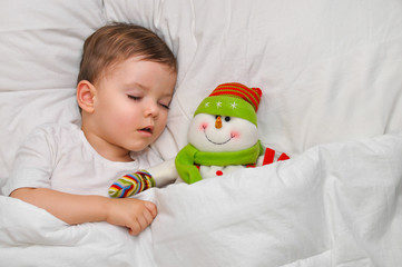 A cute toddler boy is sleeping  on white linen with his favorite toy snowman.
