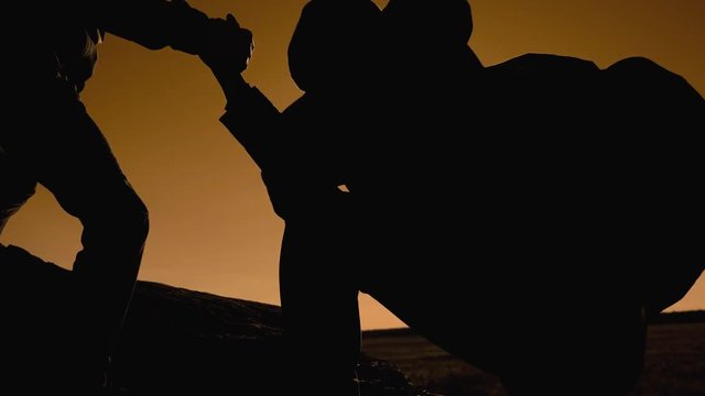 teamwork business travel trip. two men with backpacks hiking help each other silhouette in mountains with sunlight. slow motion video. teamwork friendship hiking help each other trust assistance the