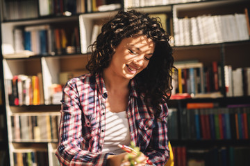 Beautiful charming young student girl sitting in library and learning. Smiling and taking notes.