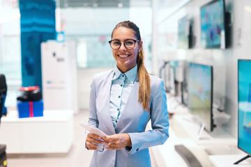 Beautiful smiling Caucasian woman dressed in suit standing in tech store and looking at camera.