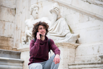Handsome young man with curly hair in tracksuit talking on his mobile phone in front of Nile God statue in Rome