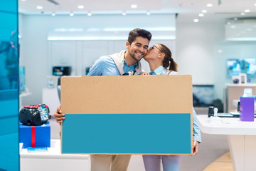 Happy multicultural couple holding unpacked television while standing in tech store. Woman kissing man in the cheek.