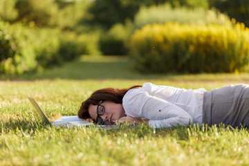 Business woman lying on a green grass and sleeping next to her laptop.