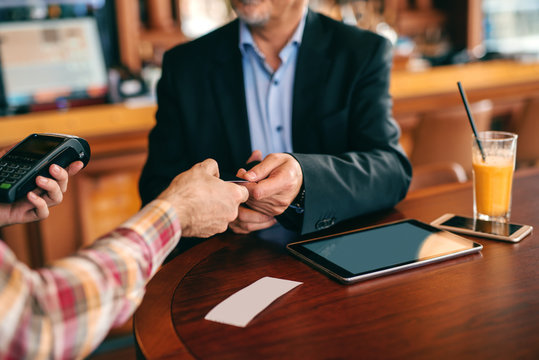 Senior adult in suit paying bill with credit card while sitting in cafeteria. On desk bill, tablet, smart phone and juice.