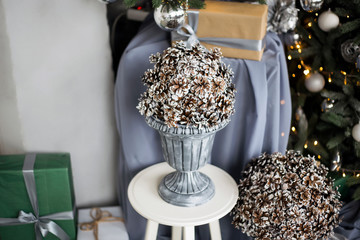 It is a lot of cones in a gray vase on a white chair against the background of a green fir-tree with a yellow garland and gifts