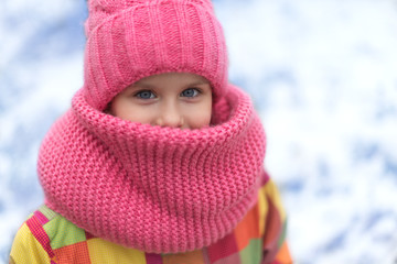 Pretty little girl with smiling eyes is looking through warm hat and snood. Cold winter weather.