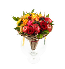Original gift in the form of a bouquet consisting of yellow and red roses, pomegranate, apples, lemon, pears wrapped in kraft paper, standing in a large glass goblet on a white background