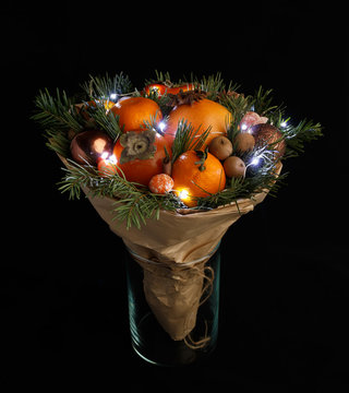 Original Christmas bouquet consisting of persimmon, mandarins, nuts, cinnamon sticks and pine twigs wrapped in kraft paper and decorated with a garland stands in a glass vase on a black background