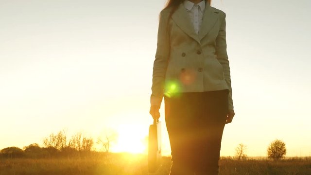 beautiful business woman in pants and jacket with briefcase in hand walks across field in rays of beautiful sunset.