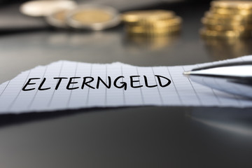 Elterngeld auf einem Zettel mit Stift vor Geldmünzen