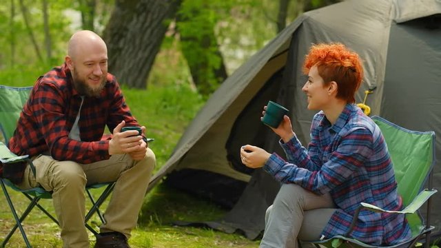 Man and woman couple talking, smiling, playing with kid outdoors near camping tent. Tourists with child travel, hiking together. Travelers enjoy nature in park, drink coffee or tea. Family holidays.