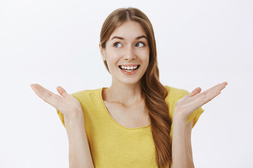 Waist-up shot of unaware carefree european female in yellow t-shirt shrugging with palms raised near shoulders gazing left and smiling with unconcerned clueless gesture over gray background