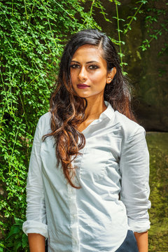 Young East Indian American Woman with long hair traveling, relaxing at Central Park, New York City, wearing white long sleeve shirt, standing by rocks with long green leaves, looking forward..
