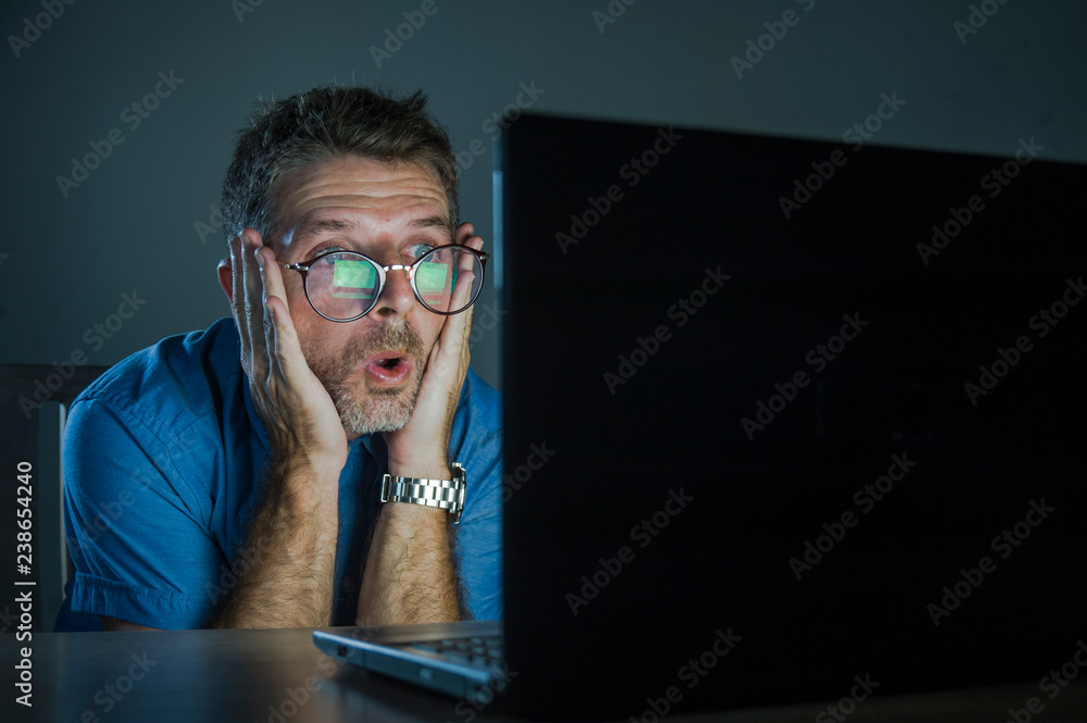 Wall mural weird shocked and surprised geek man in nerd glasses working on laptop computer late night in the da