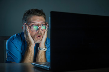 weird shocked and surprised geek man in nerd glasses working on laptop computer late night in the...