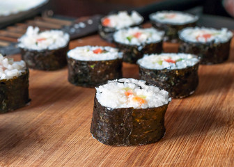 Chumaki Sushi Rolls with Tuna, Salmon, Rice, Cucumber, Avocado And Nori Seaweed on a Wooden Board with Kitchenware Blurred in Background.