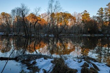 The last sliver of sunshine falls upon the trees across the pond with mirror reflections on the surface and patchy remnants of snow on the ground at Yates Mill County Park in Raleigh North Carolina.