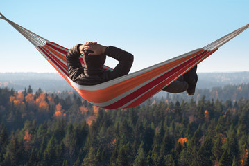 A man is lying on a hammock on a hill above a pine forest. His hands behind his head.