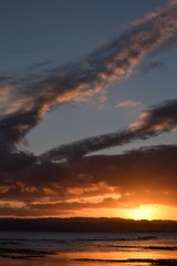 The orange sun sets beneath the odd shaped clouds above the beach in Gisborne, New Zealand.