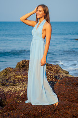 full length portrait of traveller woman in blue chiffon dress enjoys her tropical beach vacation standing on a rock indian ocean waves and sky background.attractive girl wear summer collection outfit