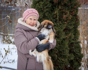 Portrait of an elderly woman with a dog in her arms. Lady of plus size with little red senior pekingese dog. Woman with dog in winter village.