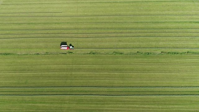 Aerial high altitude footage of tractor pulling loader wagon collecting dried grass piled up in straight lines beautiful agricultural scene looking down on green pastures 4k high resolution quality