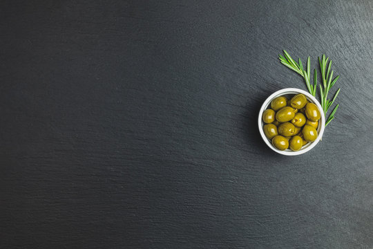 Set of green olives on plate and rosemary on a black stone background