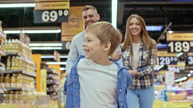 Cute little boy smiling, pulling shopping cart and walking towards the camera through supermarket aisle while mother and dad going behind him