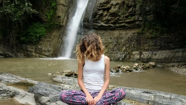 Woman is sitting in the lotus position and doing exercises on the background of a large waterfall